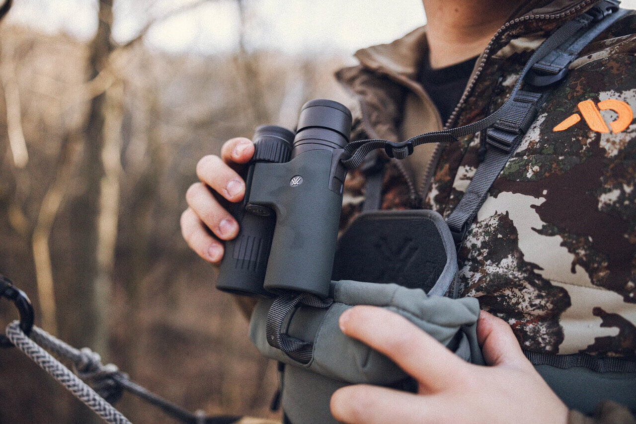 Person hält ein grünes Fernglas vor einem Waldhintergrund im Freien.