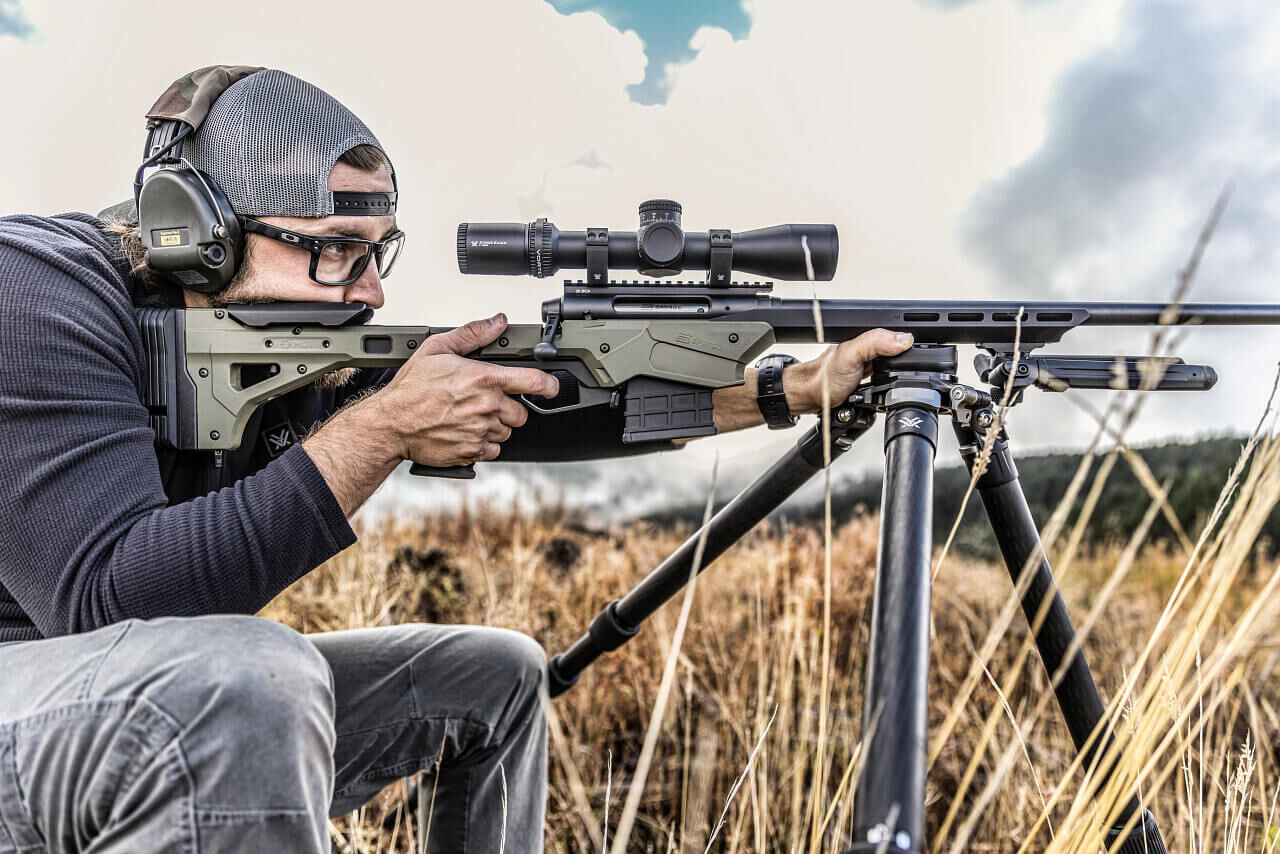 Mann mit Gewehr auf einem Stativ im Grasland, trägt Schutzbrille und Gehörschutz
