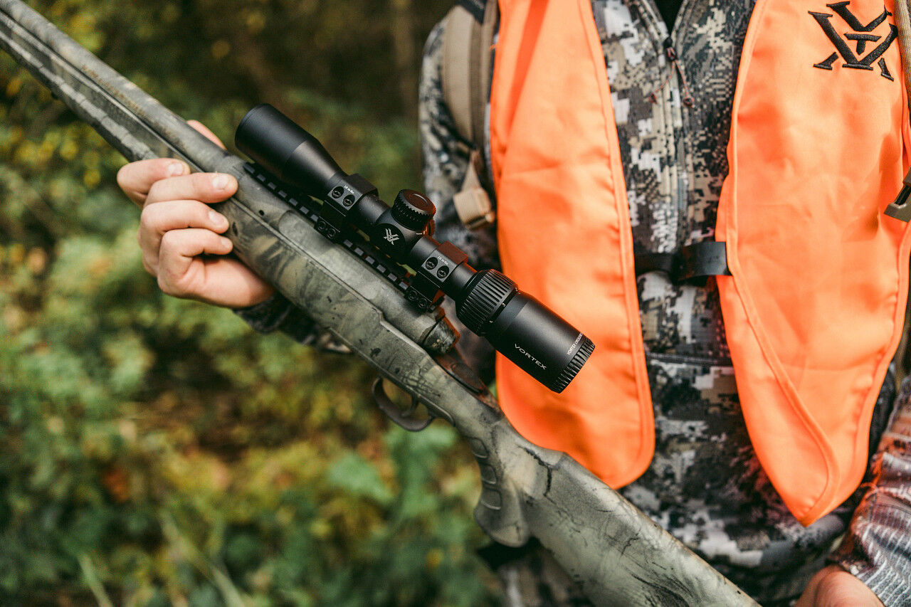 Ein Jäger in oranger Sicherheitsweste und Tarnkleidung hält ein Gewehr mit einem montierten Zielfernrohr in der Hand. Die Szene spielt im Grünen, möglicherweise im Wald.