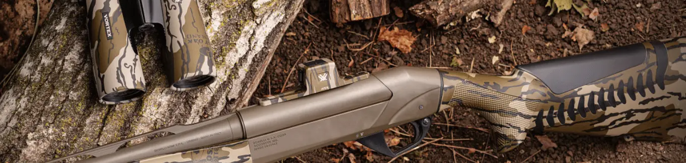 Detailaufnahme einer jagdlichen Camouflage-Flinte mit Reflexvisier neben passendem Fernglas im Tarnmuster auf Baumstamm im Wald