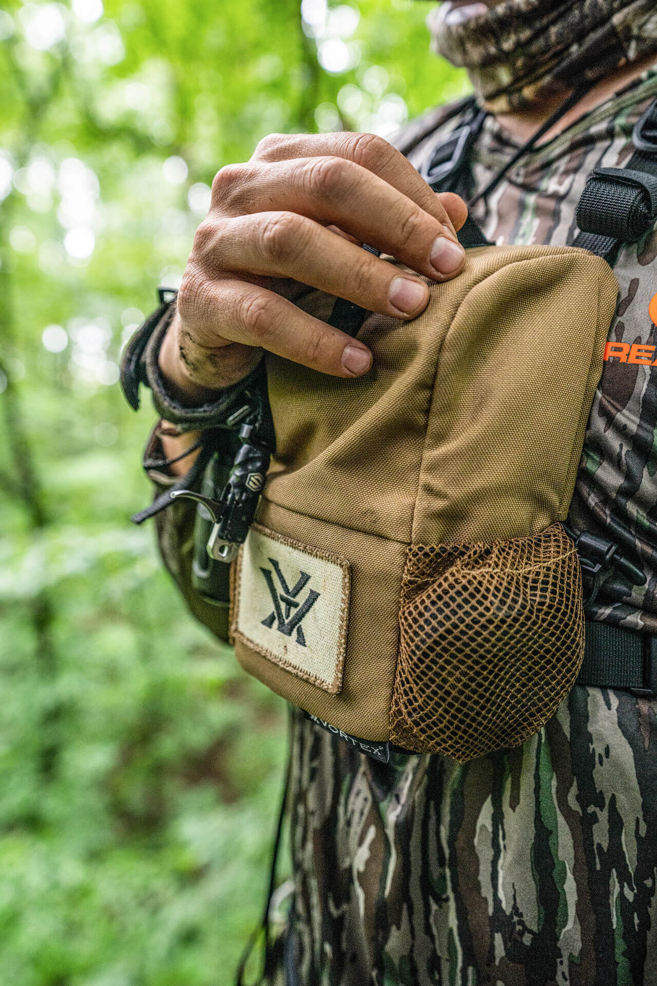 Nahaufnahme eines Mannes in Tarnkleidung, der eine braune kleine Tasche mit Mesh-Seitentasche in einem Wald hält