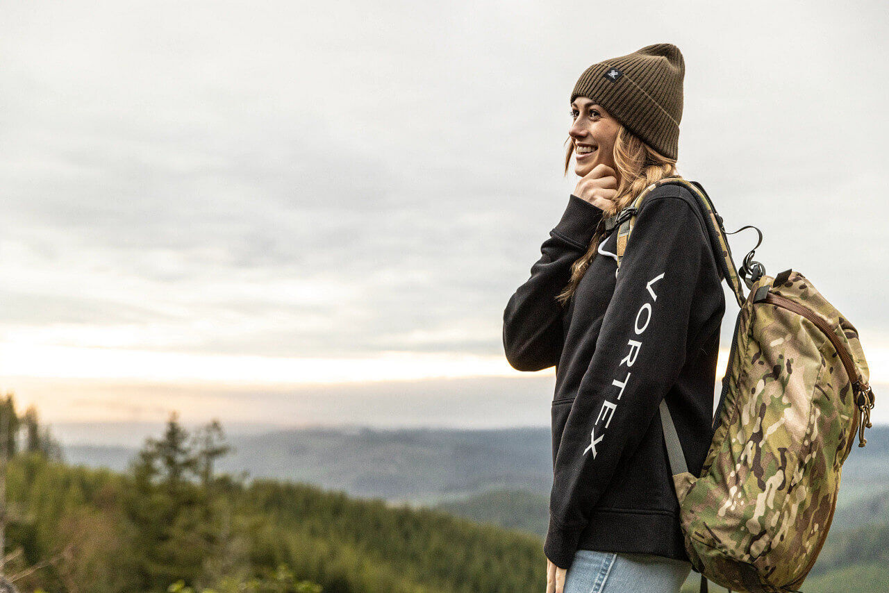 Eine lächelnde Frau in Outdoor-Kleidung mit Rucksack und Mütze, vor einer malerischen Landschaft mit Wald und Himmel im Hintergrund.